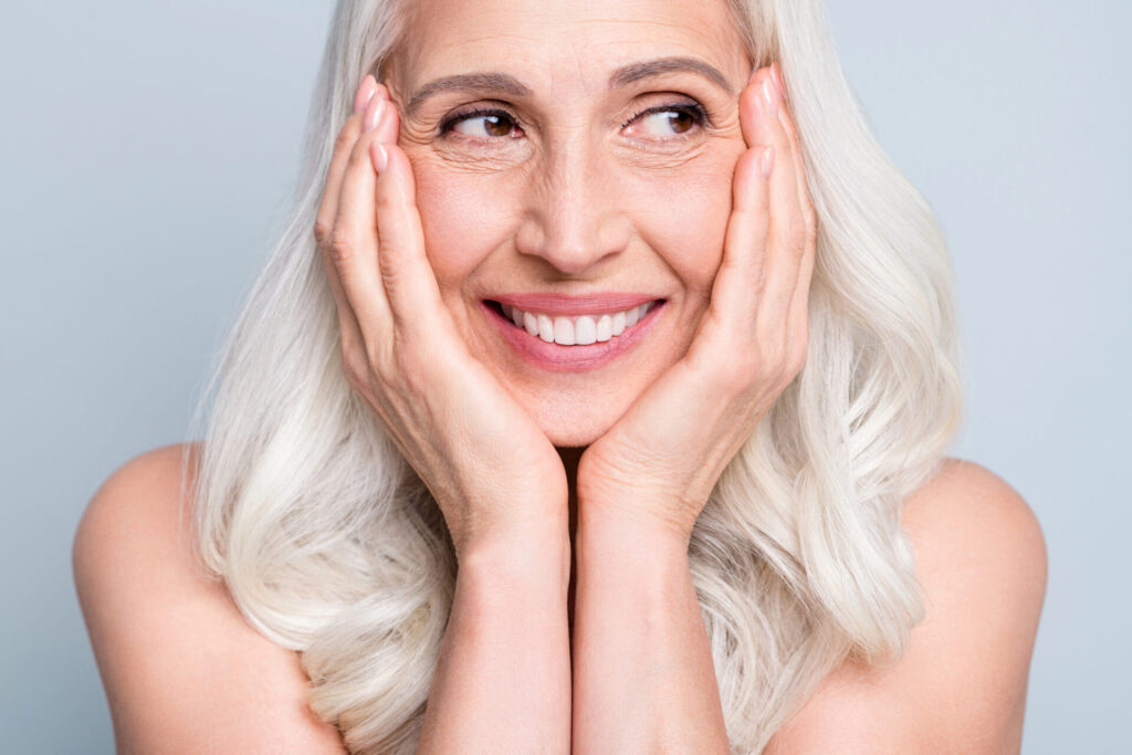 Starsza kobieta szeroko się uśmiecha i patrzy w lewo, opierając twarz o dłonie