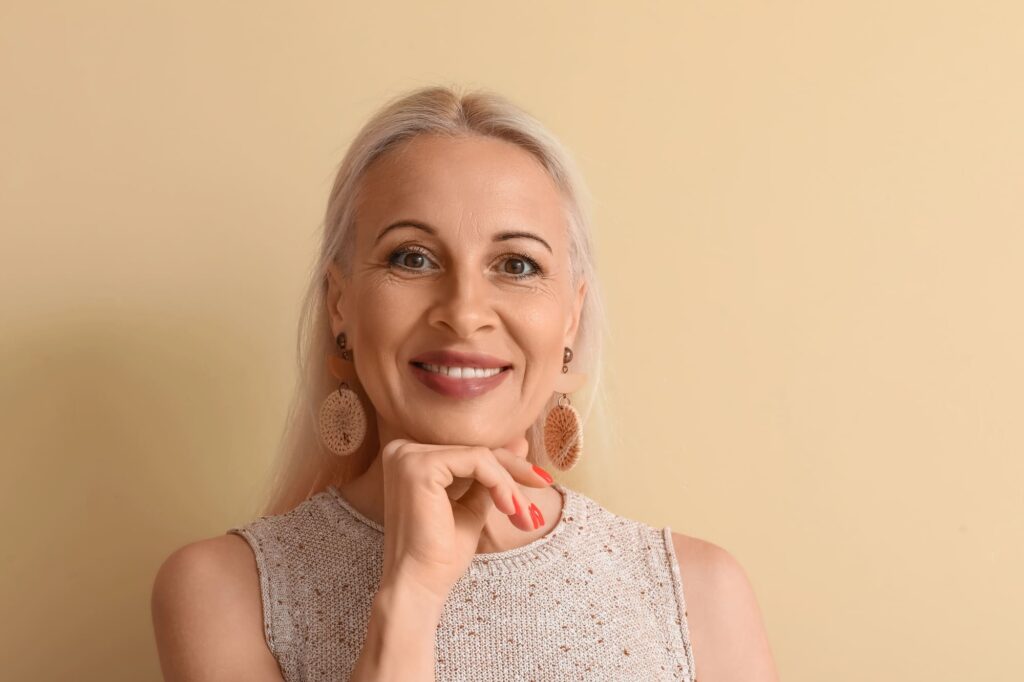 Kobieta na kremowym tle uśmiecha się do zdjęcia, podpierając podbródek dłonią